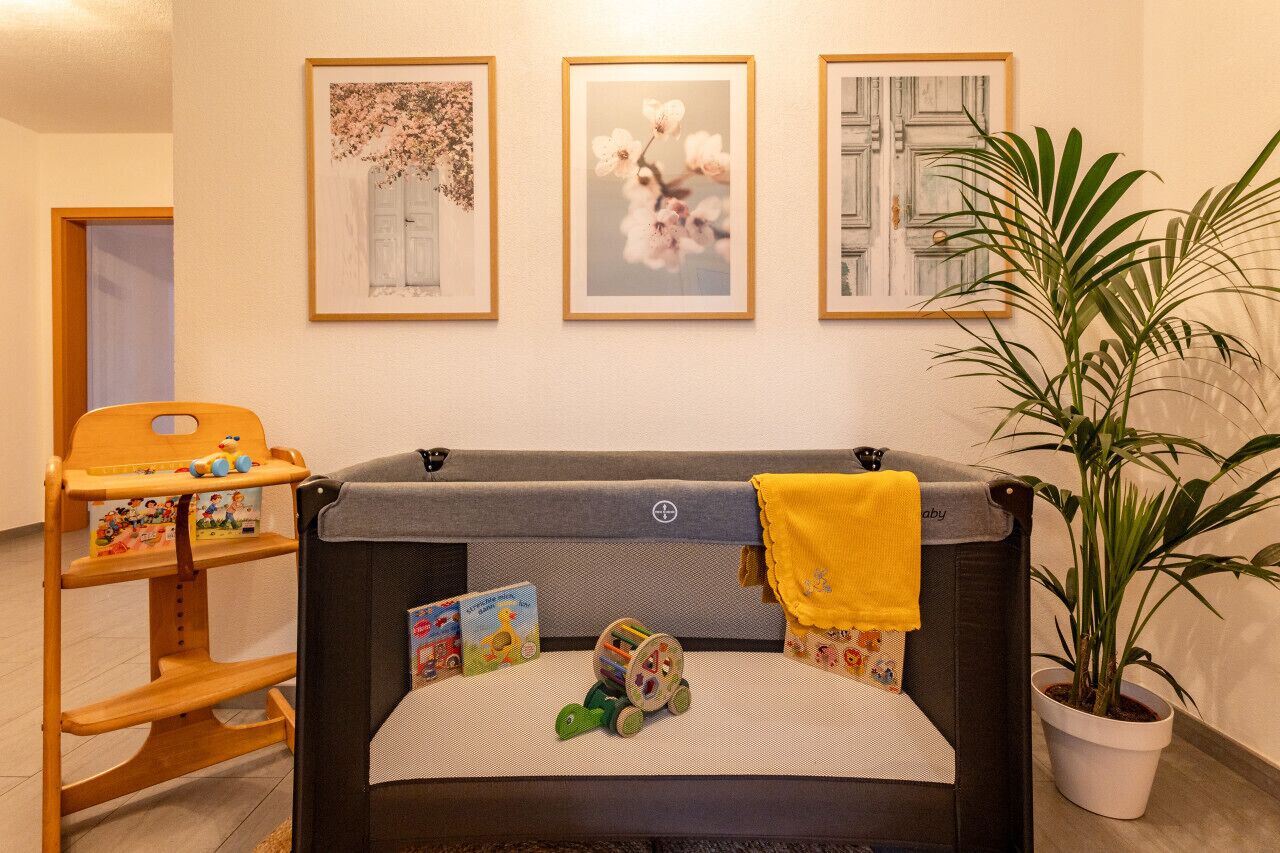 Un espace pour changer les bébés, équipé d’un siège pour bébé, de jouets et de livres. Des tableaux encadrés ornent le mur, et une plante en pot est également présente.