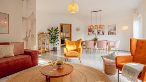 Un salon moderne avec un canapé rouge, des fauteuils orange, une table basse ronde et un coin repas comprenant une table blanche et des chaises roses.
