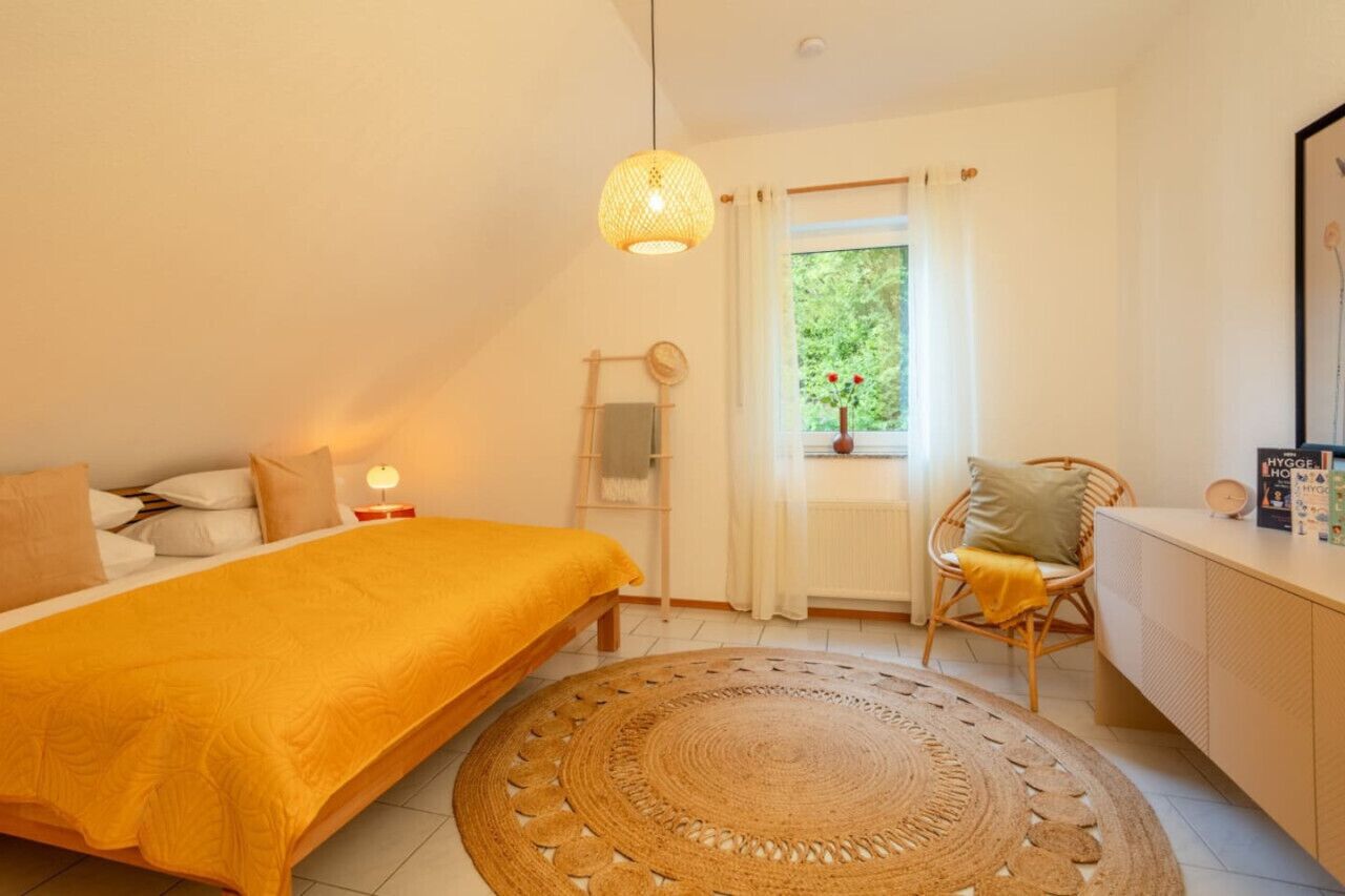 Une chambre à coucher comprenant un lit, une table de chevet, une chaise, une fenêtre avec des rideaux et un luminaire suspendu.