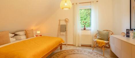 Une chambre à coucher comprenant un lit, une table de chevet, une chaise, une fenêtre avec des rideaux et un luminaire suspendu.