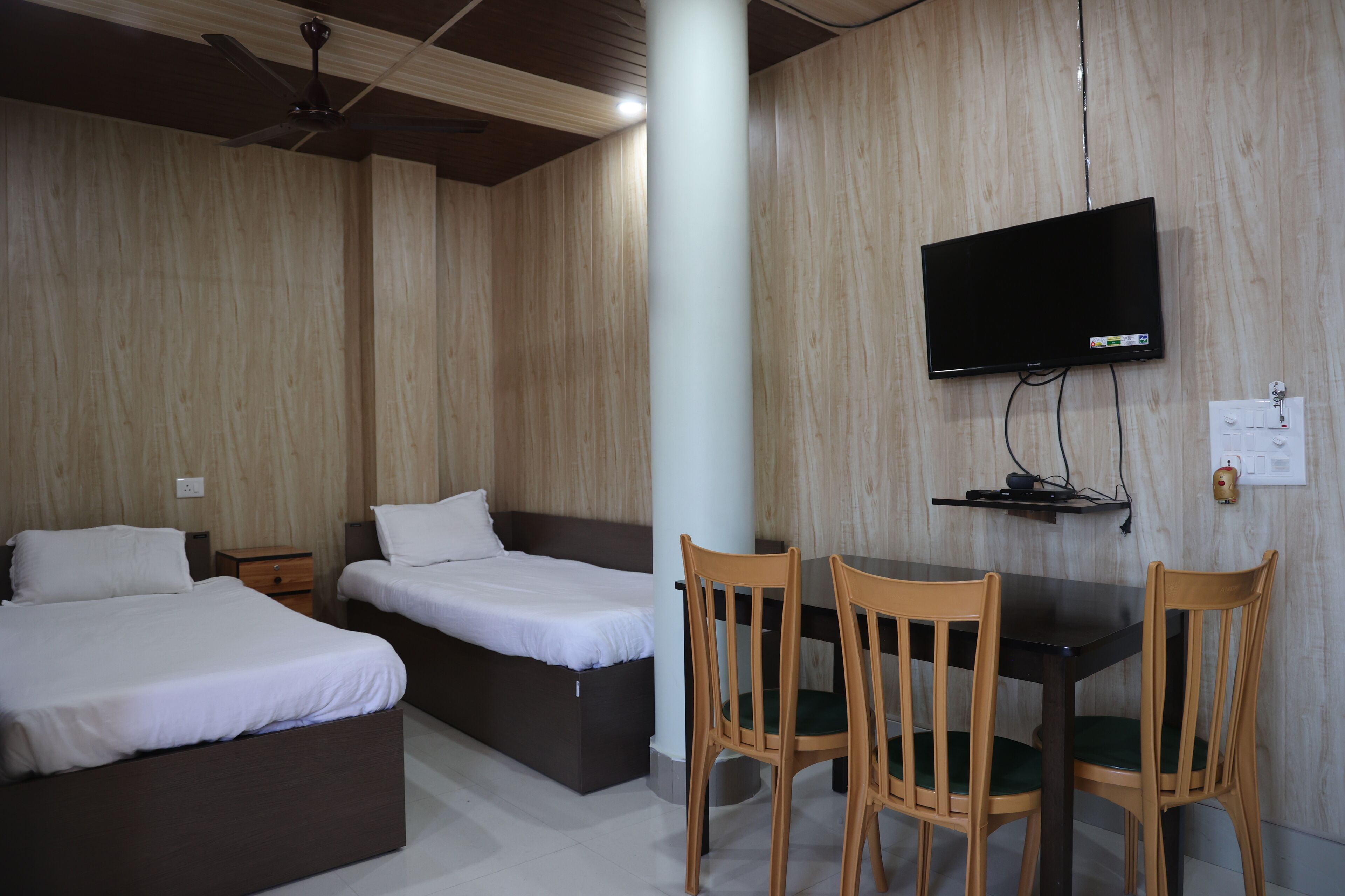 Una habitación con dos camas, una mesa con sillas, un televisor instalado en la pared y un ventilador de techo.