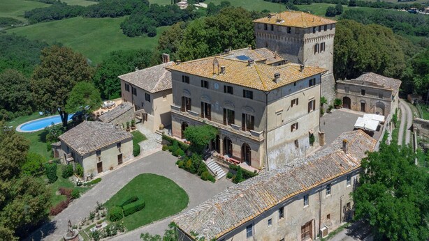 A large, historic building with multiple floors and a tower, surrounded by lush greenery and a swimming pool.