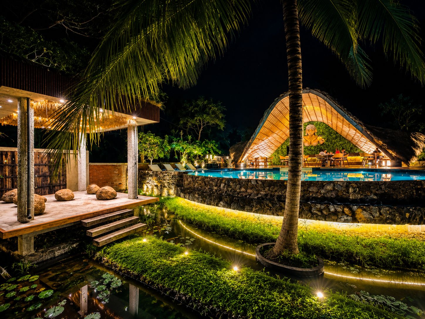 A nighttime resort with a swimming pool, outdoor seating, and a thatched-roof structure.