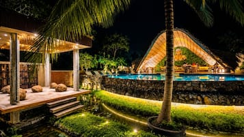 A nighttime resort with a swimming pool, outdoor seating, and a thatched-roof structure.