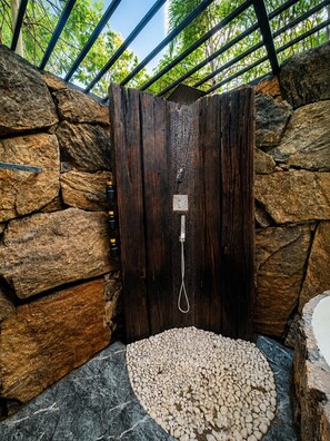 A stone-walled shower area with a wooden door, a pebbled floor, and a metal showerhead. - Woodland Sanctuary (Habarana)