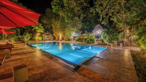 Una piscina con solárium de azulejos, rodeada de árboles y un pequeño edificio.