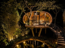 Una estructura circular de madera con una escalera que lleva a un balcón, rodeada de exuberante follaje y un pequeño estanque.