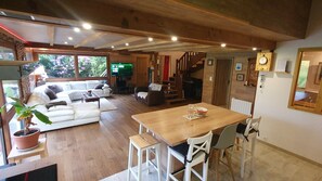 A cozy living area with a wooden dining table, white chairs, a sofa, and a staircase leading to another room. - Le chalet breton, Enjoy your vacations in a warm and green setting (loperhet)