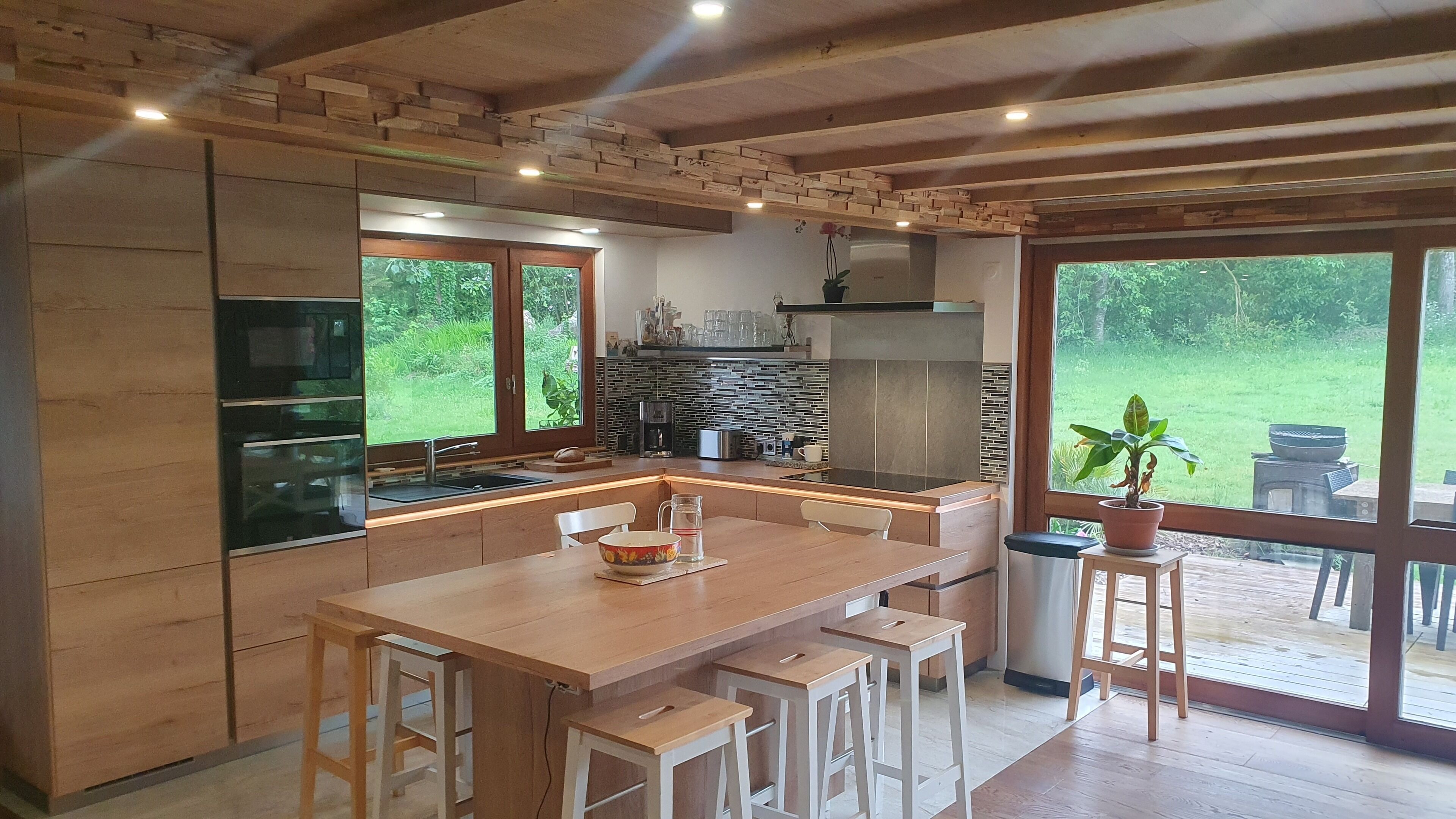 A modern kitchen with wooden cabinetry, a central island with bar stools, a coffee machine, and a view of the outdoors.