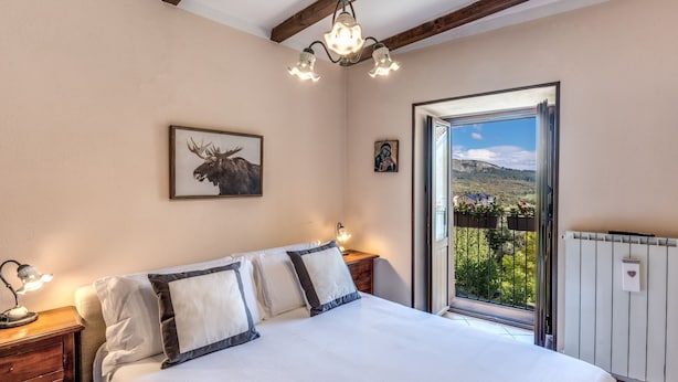 Une chambre à coucher comprenant un lit, une table de chevet, une lampe, un tableau encadré représentant un élan, une icône religieuse et un balcon offrant une vue sur la verdure et les bâtiments.