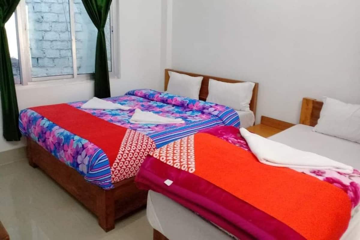 Uma cama de casal com colcha floral e manta vermelha, uma cama simples com lençol branco e uma janela com cortinas verdes.