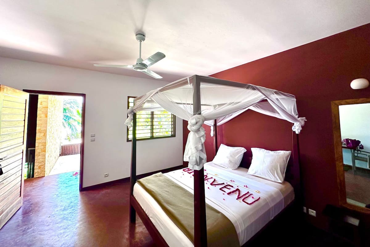 A room with a four-poster bed, a ceiling fan, and a window with bars.
