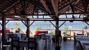 An outdoor dining area with wooden tables and chairs, potted plants, and a view of the beach and ocean. - ANKI LODGE (Nosy Be)