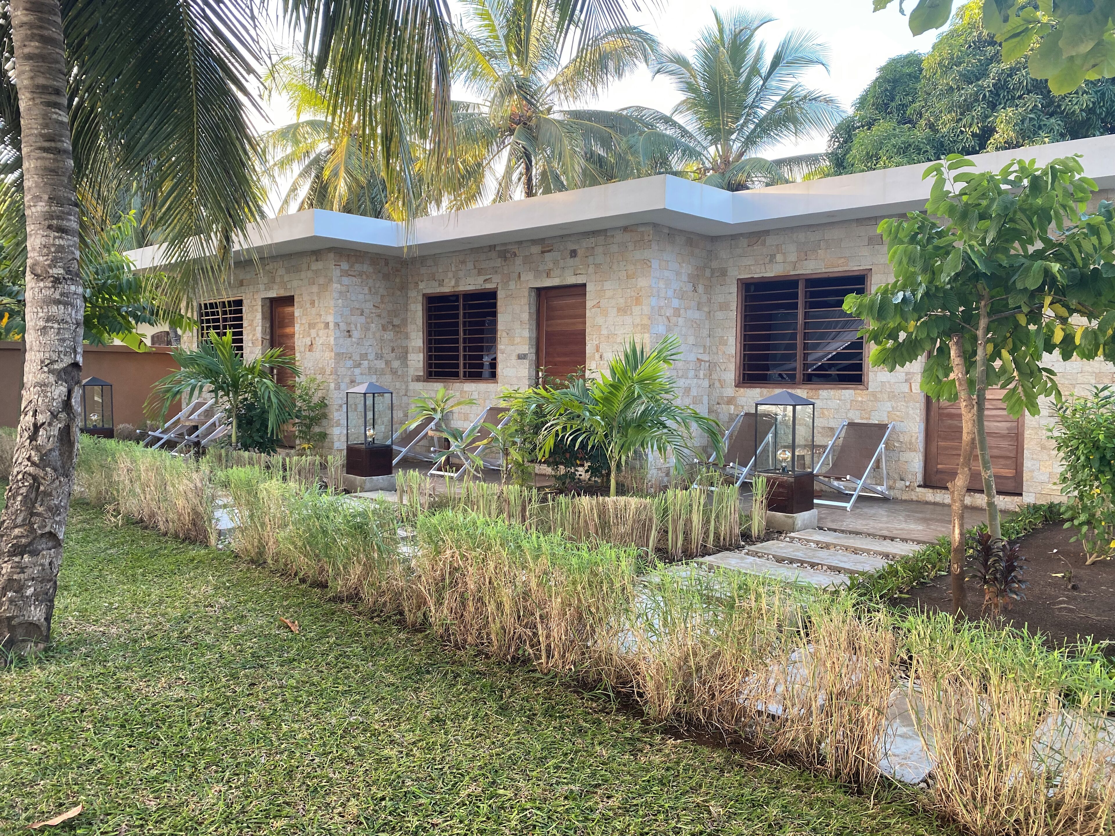 A building with a stone facade, multiple windows, and a garden area with grass, palm trees, and lounge chairs.