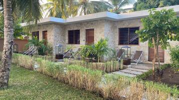 A building with a stone facade, multiple windows, and a garden area with grass, palm trees, and lounge chairs.