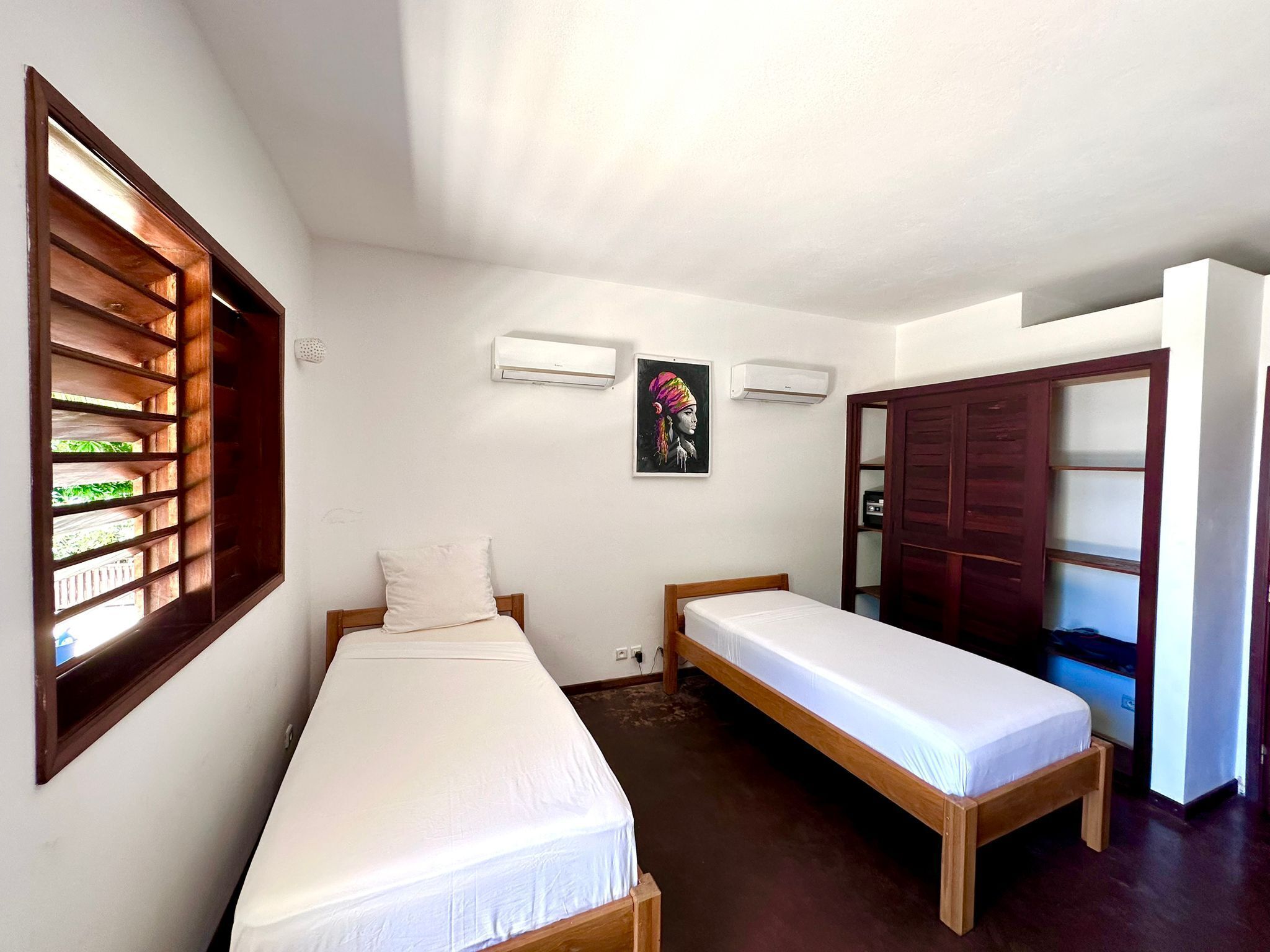 A room with two single beds, a window with wooden shutters, an air conditioning unit, and a door leading to another room.