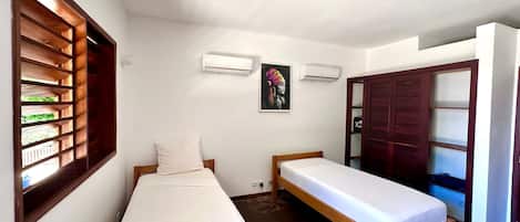 A room with two single beds, a window with wooden shutters, an air conditioning unit, and a door leading to another room.