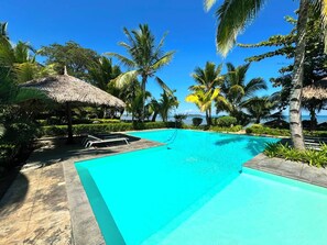 A clear blue pool with lounge chairs and a thatched umbrella, surrounded by palm trees and a view of the ocean. - ANKI LODGE (Nosy Be)