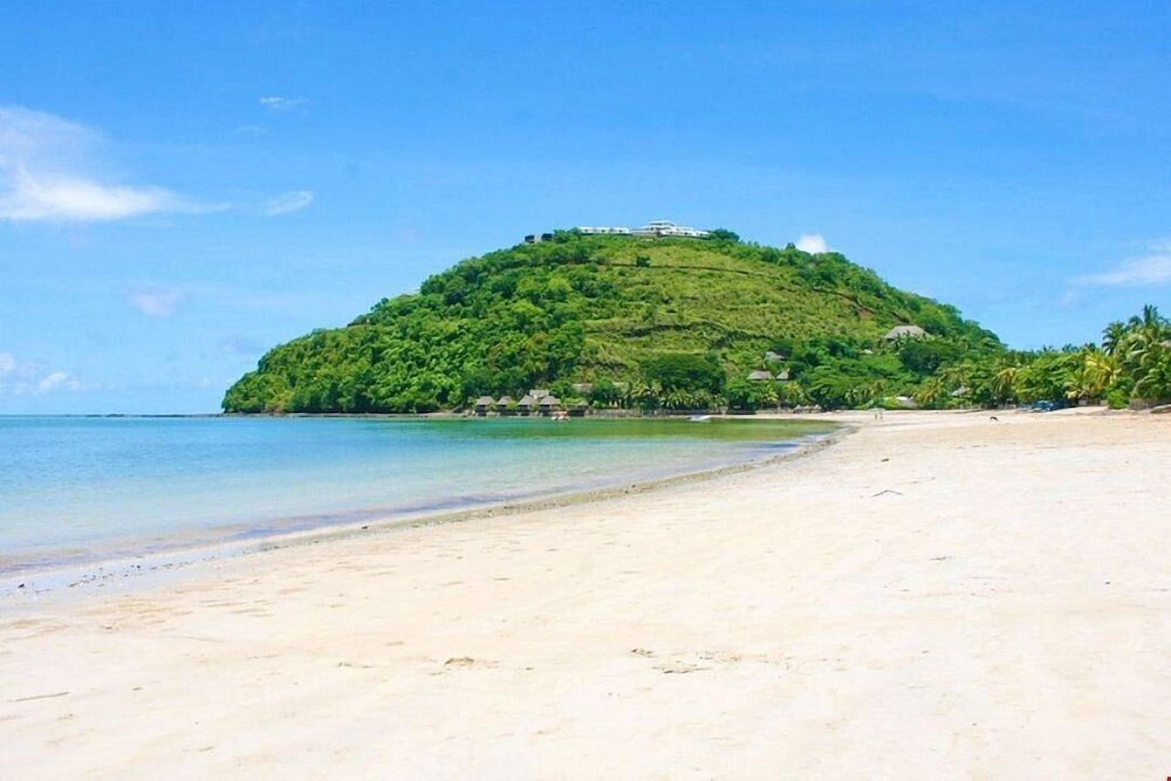 A beach with a hill in the background, clear blue water, and a few buildings on the hill.