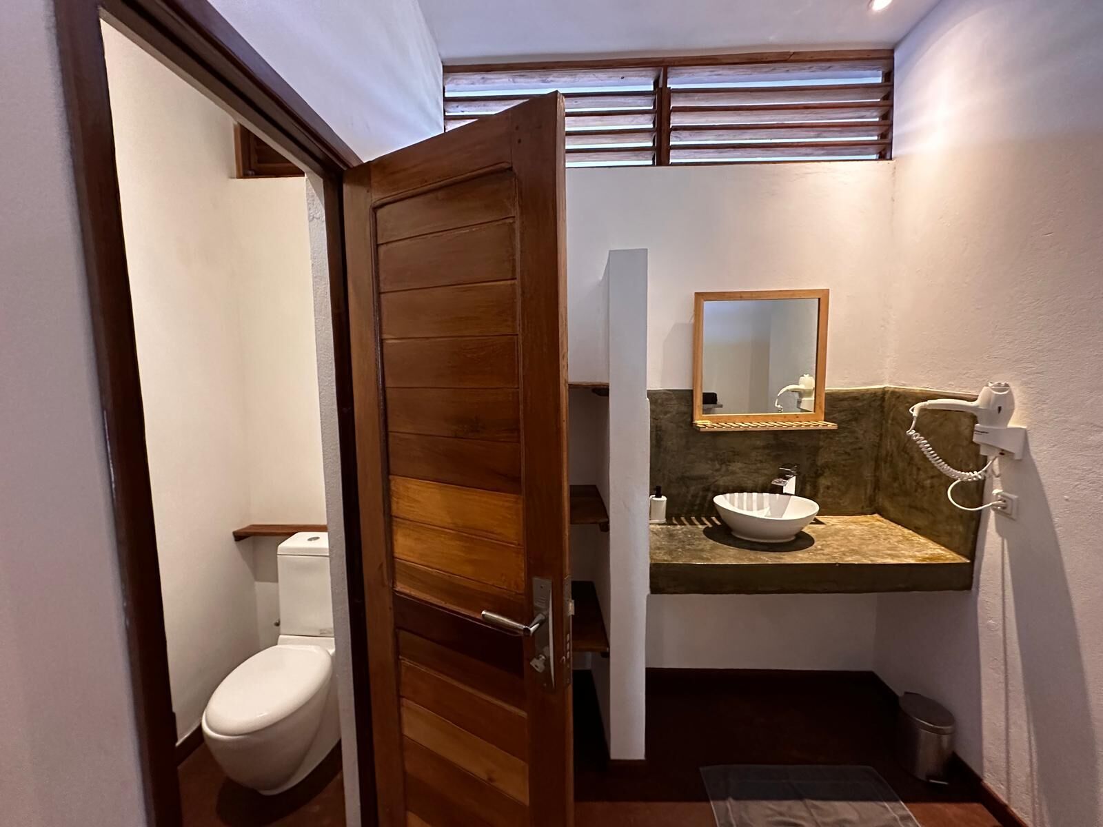 A compact bathroom with a wooden door, a white toilet, a sink with a bowl, a mirror, and a hairdryer on the wall.