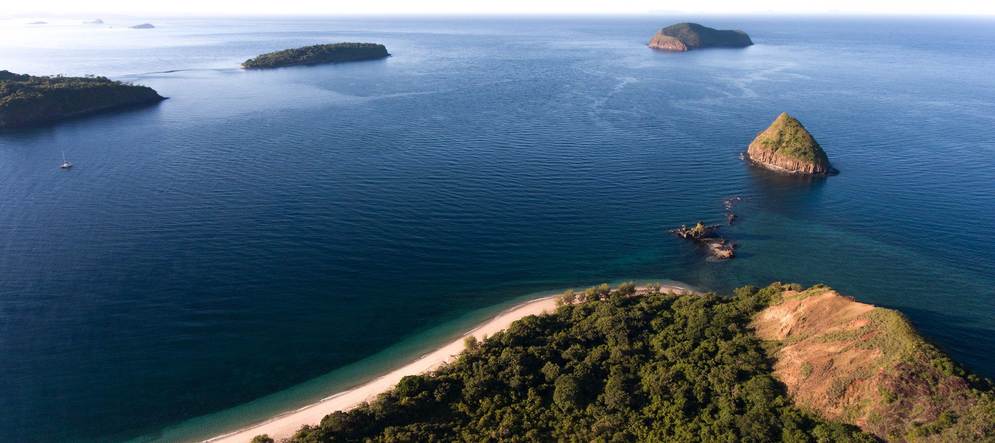 An aerial view of a coastal area with a sandy beach, dense greenery, and several small islands in the distance.
