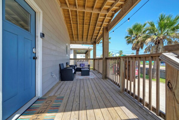 Una terraza de madera con mobiliario exterior, una puerta azul y vistas a la calle y palmeras.