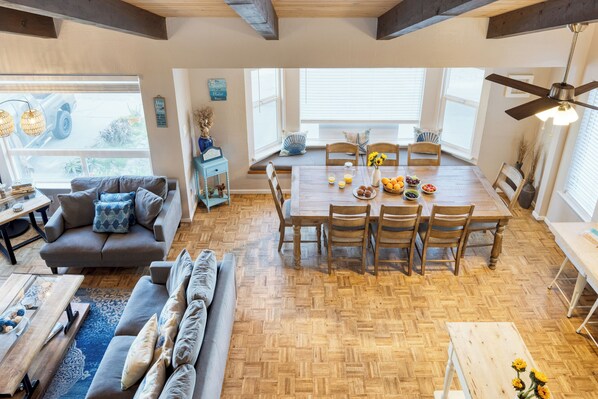 Un vaste espace de vie doté d’un parquet en bois disposé en point de Hongrie, d’une table à manger pour six personnes et d’une grande fenêtre avec des stores.