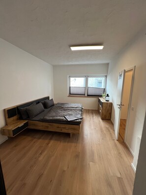A bedroom with a wooden bed, a window with blinds, a bedside table, and a door.