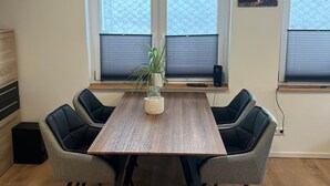 A dining area with a wooden table, four grey upholstered chairs, a potted plant, and two windows with blinds.