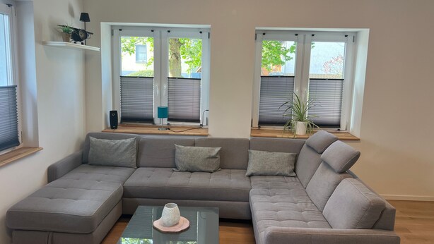A modern living room with a sectional sofa, a glass coffee table, and two windows with blinds.