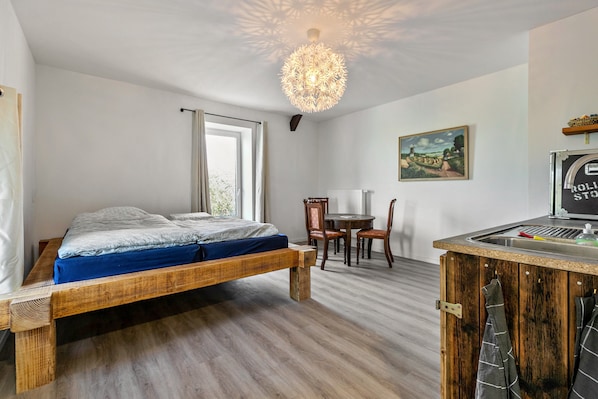 A bedroom with a wooden bed, a dining table with chairs, a kitchen area with a sink, and a ceiling light fixture.