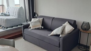 Un salon moderne avec un canapé gris, deux coussins, une petite table d’appoint avec une lampe et une bibliothèque.