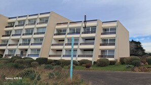 Un immeuble résidentiel à plusieurs étages, doté d’une architecture moderne, avec des balcons et un jardin paysager à l’avant, agrémenté d’arbustes et de rochers.