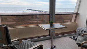 Un balcon avec vue sur la plage et la jetée, une petite table avec une plante et deux chaises.