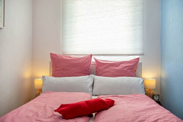 A bed with red and white checkered bedding, two pillows, and a red throw. There are two bedside lamps and a clock on the nightstand.