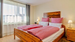 Una cama doble con ropa de cama roja y blanca, un cabecero de madera, una mesita de noche con lámpara y una ventana con cortinas translúcidas.