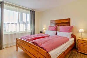 Una cama doble con ropa de cama roja y blanca, un cabecero de madera, una mesita de noche con lámpara y una ventana con cortinas translúcidas.