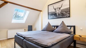 Een slaapkamer met een bed, een raam, een radiator, een aan de muur hangend schilderij en een nachtkastje.