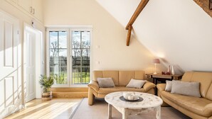 Een moderne woonkamer met een beige bank, een ronde salontafel en een groot raam met uitzicht op bomen.