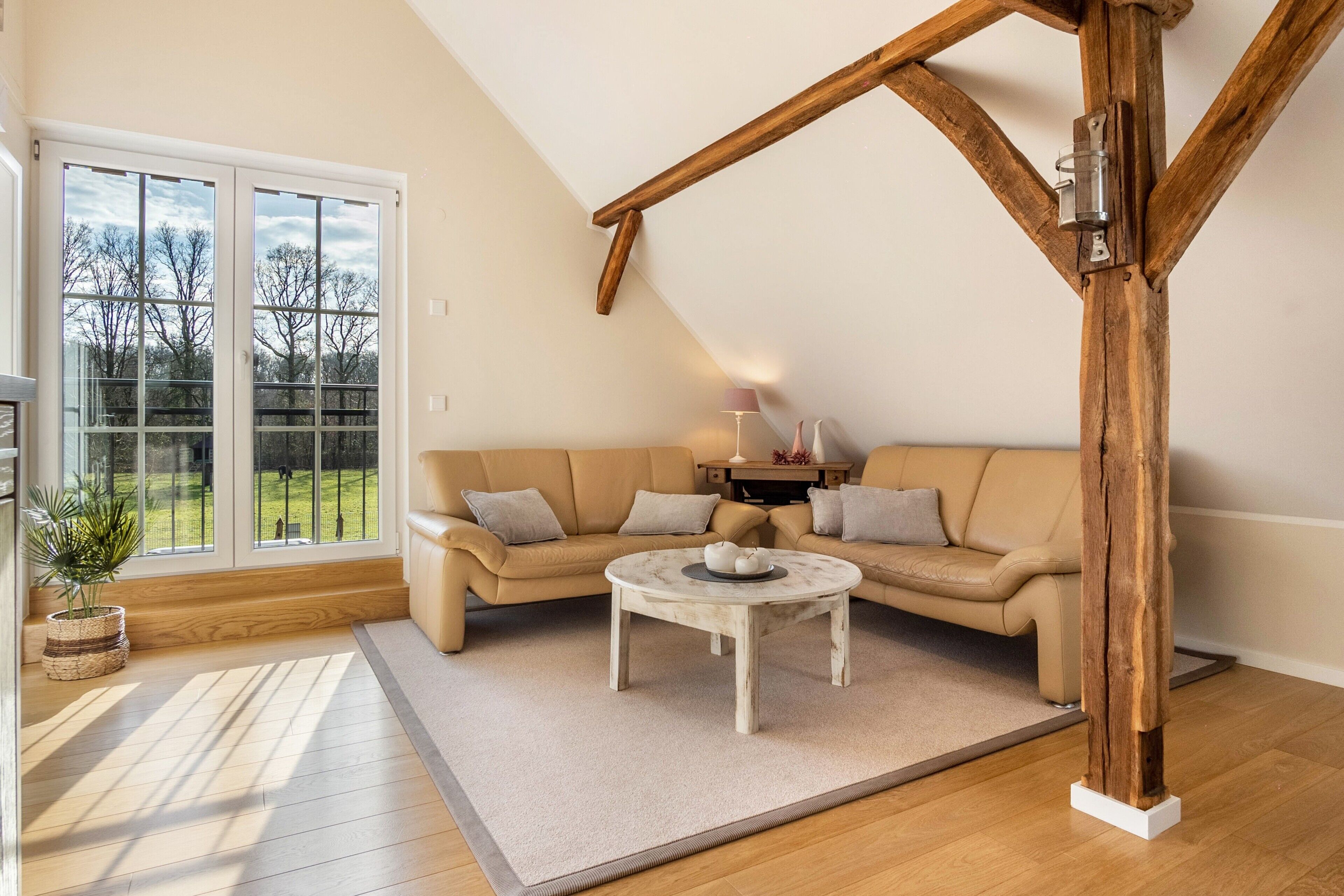 Ein Zimmer mit Holzbalkendecke, einem beigen Sofa, einem kleinen runden Tisch und einem großen Fenster mit Blick auf Bäume.