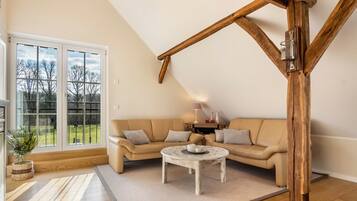 Ein Zimmer mit Holzbalkendecke, einem beigen Sofa, einem kleinen runden Tisch und einem großen Fenster mit Blick auf Bäume.