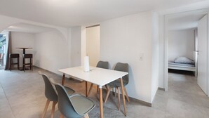 Ein helles, modernes Zimmer mit einem weißen Tisch, vier Stühlen und einem Bett im Hintergrund.