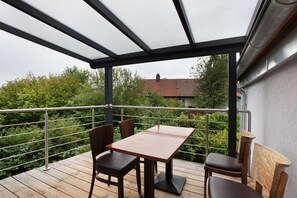 A balcony with a table and chairs, a metal railing, and a view of greenery and a building in the background.