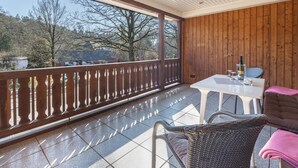 Un balcón techado con vistas a árboles y casas, una mesa blanca, sillas de mimbre y una barandilla de madera.
