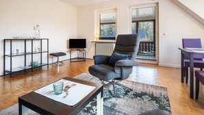 Una sala de estar moderna con un sillón gris oscuro, una mesa de centro negra, un aparador de vidrio, un televisor de pantalla plana y vistas al exterior a través de una ventana.