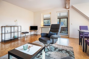 Un salon moderne avec un fauteuil gris foncé, une table basse noire, une vitrine, un téléviseur à écran plat et une vue sur l’extérieur par une fenêtre.