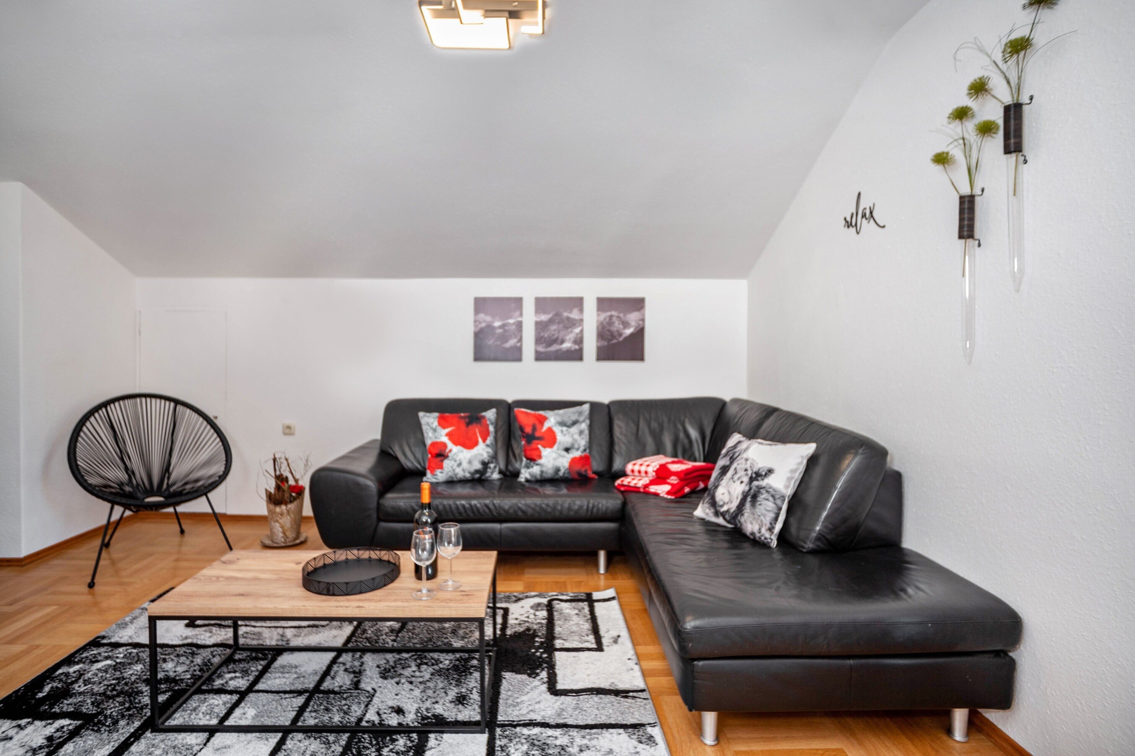 Una sala de estar moderna con un sofá de cuero negro, una mesa de centro de madera y una silla negra. Hay tres cuadros enmarcados en la pared y un florero con flores a la derecha.