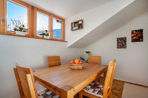 A dining room with a wooden table, chairs, a fruit basket, and a window with a view of mountains.