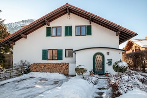 A two-story building with a sloped roof, a green door, and a stone wall with stacked firewood.
