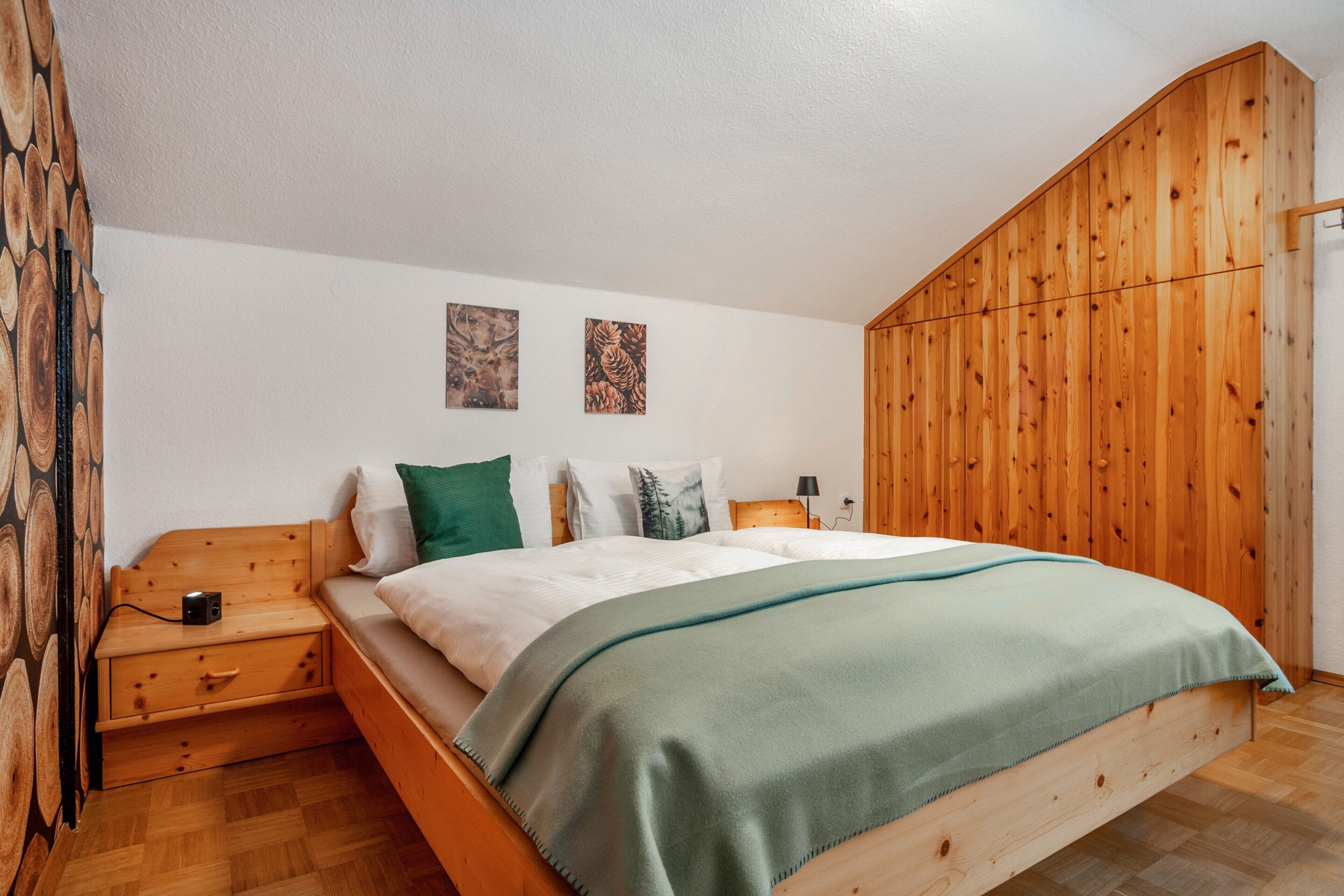 Ein hölzernes Bett mit grüner Decke, ein Nachttisch mit Lampe und zwei an der Wand hängende Bilder.